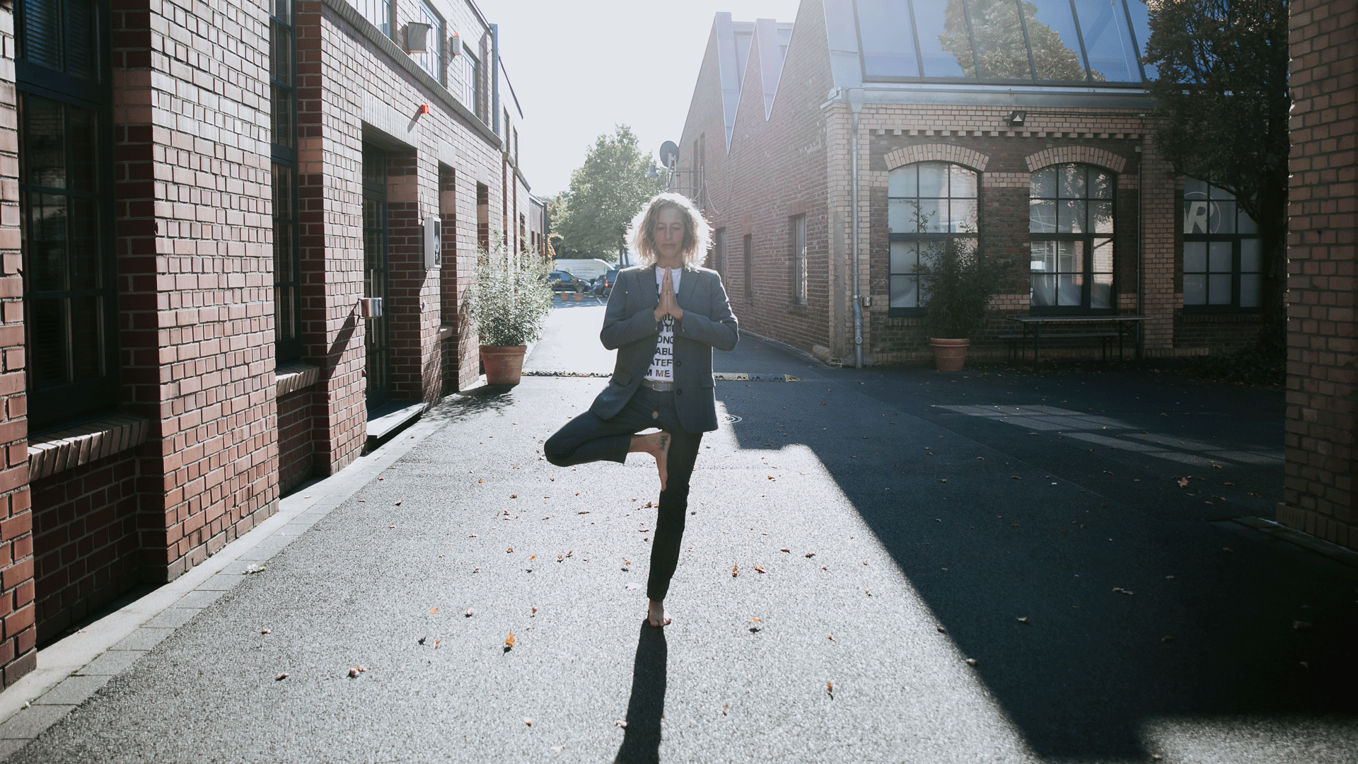 Bianca steht im Business-Look barfuß auf der Straße und macht die Pose "Der Baum".